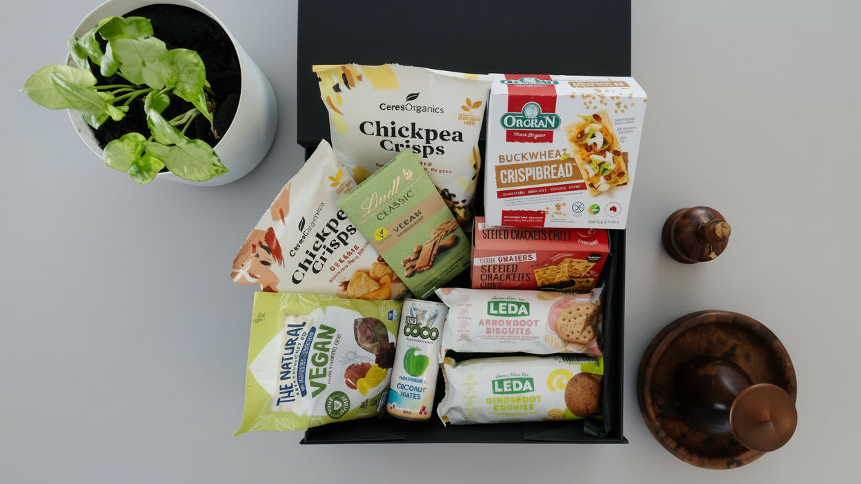 #VeganTreats#ArchiesGiftBox A vegan treat gift box containing various packaged foods such as chickpea crisps, biscuits, cookies, coconut water, and more, displayed on a table with a plant in the background.