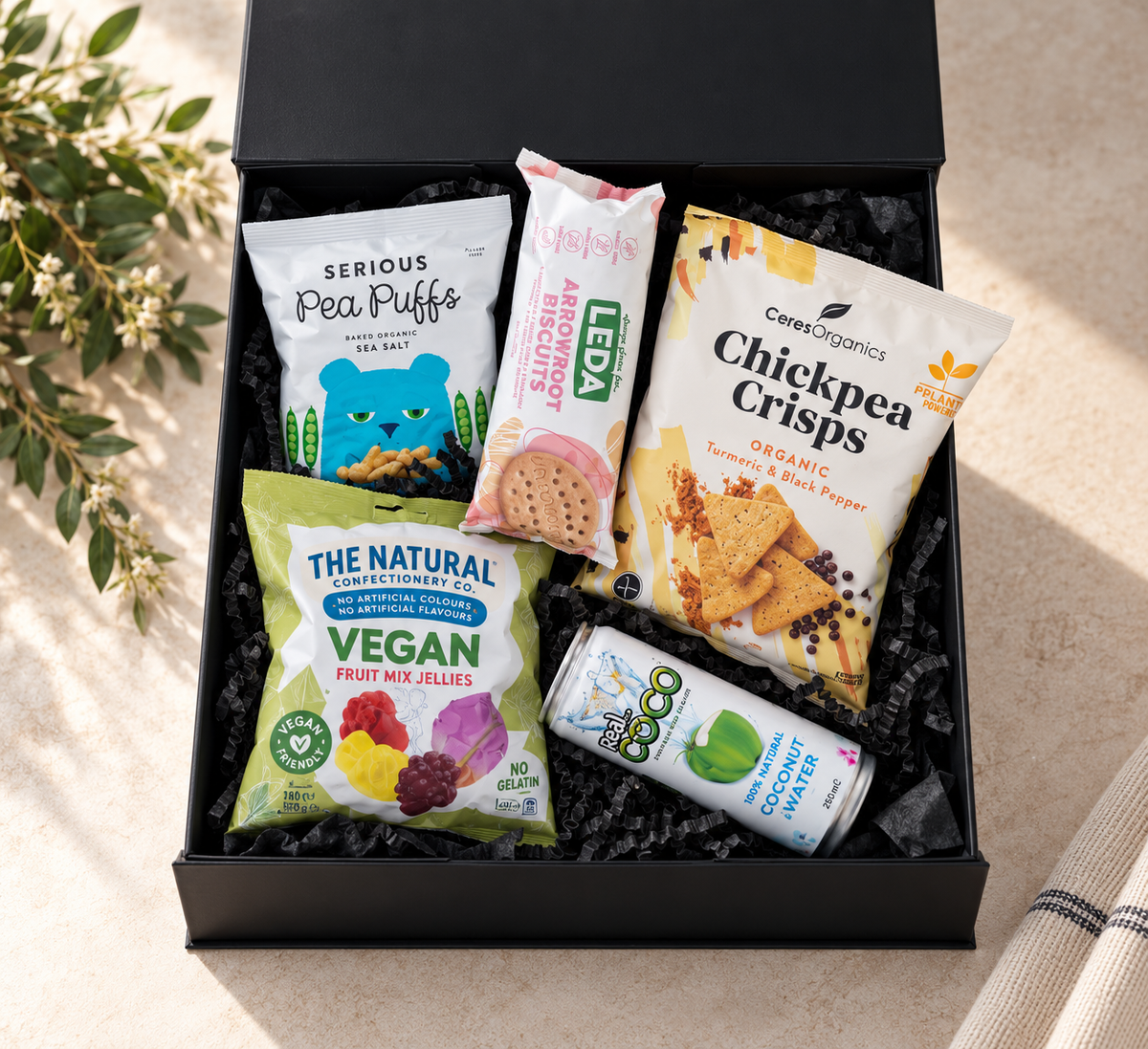 A gift box with various plant-based snacks including vegan jellies, chickpea crisps, arrowroot biscuits, pea puffs, and coconut water. Hamper Box