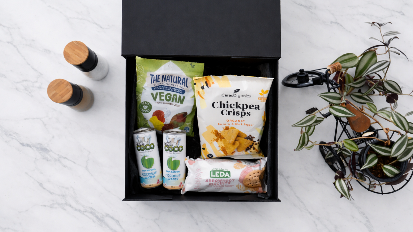 Black gift box with vegan snacks on a white surface with a plant and bottles in the background Gift-VeganGoodies-ArchiesGiftBox