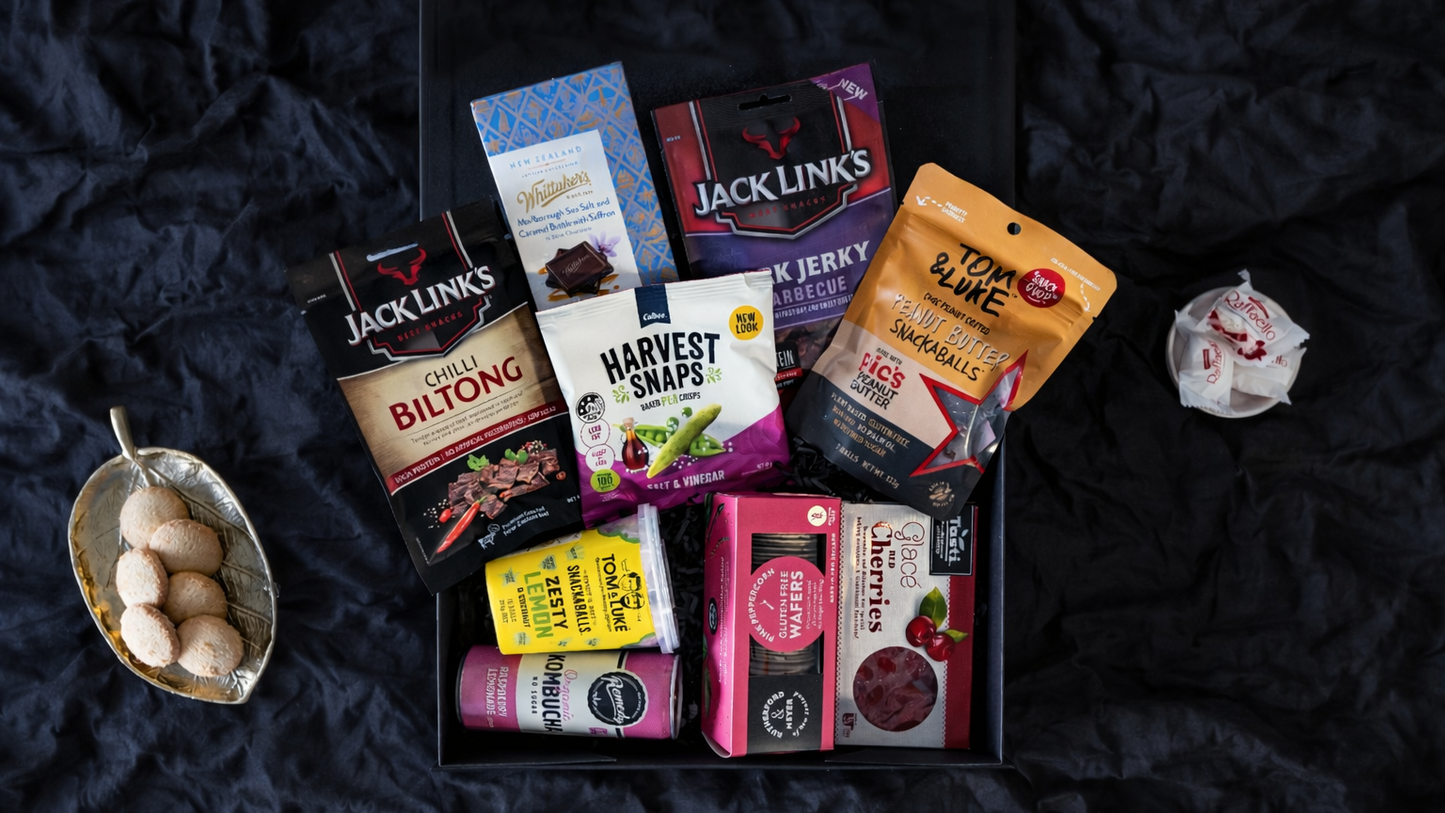 Assorted snack packages on a dark textured surface. Gifts-ChicCollection-ArchiesGiftBox