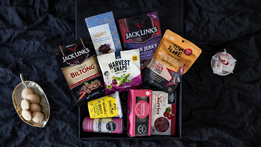 Assorted snack packages on a dark textured surface. Gifts-ChicCollection-ArchiesGiftBox